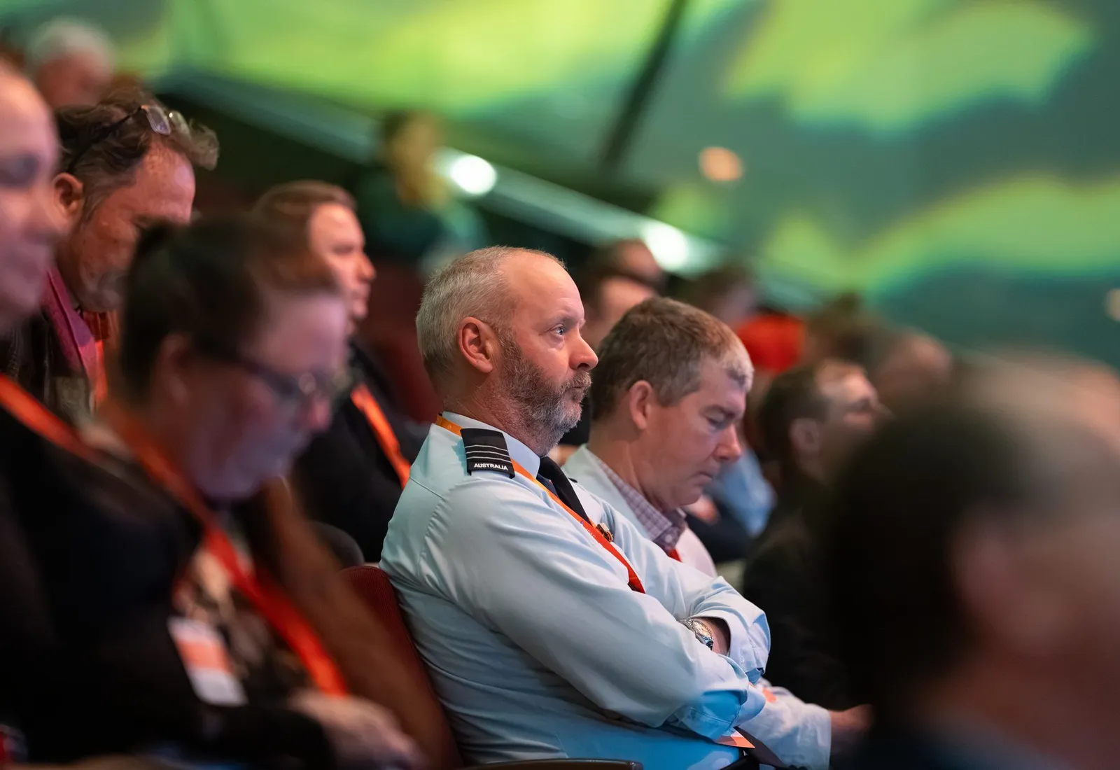 Delegates in ADSTAR lanyards watching a plenary session, the stage lighting washing the hall green