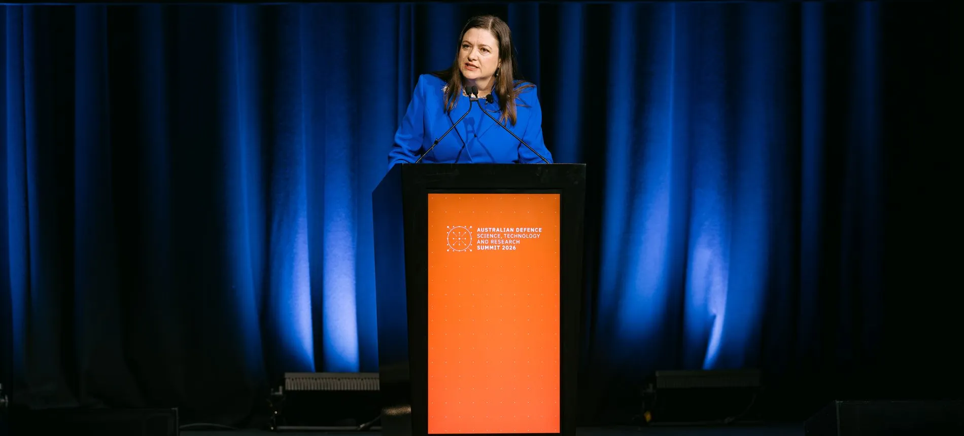 A speaker at the ADSTAR 2026 lectern, lit against blue stage drapes