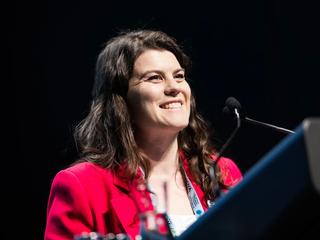 Speaker in a red jacket at a conference lectern, smiling mid-presentation